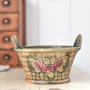 Könnte beinhalten: Ein Keramikkorb mit zwei Griffen, verziert mit einem geflochtenen Muster und drei rosa Rosen mit grünen Blättern.