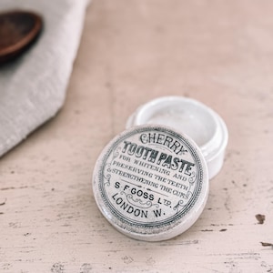May include: A vintage white ceramic jar of Cherry Tooth Paste with the lid off. The lid has black text that reads "Cherry Tooth Paste" and "S.F. Goss Ltd. London W.". The jar is on a light-colored wooden surface.