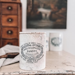 May include: A vintage, cylindrical marmalade jar with a distressed, crackled white finish. The jar features black text that reads "JAMES KEILLER & SON'S DUNDEE MARMALADE" and "GRAND MEDAL OF MERIT VIENNA 1873". A wooden spoon and a painting are in the background.