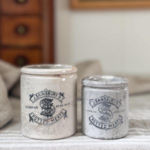 May include: Two cylindrical ceramic pots with a cream-colored crackle glaze finish. Each pot features a black printed label reading J. Sainsbury's Superior Home Made Potted Meats with a central letter S design. The pots vary in height and width.