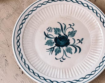 May include: A white ceramic plate with a blue and green floral design. The plate has a raised rim and a scalloped edge.