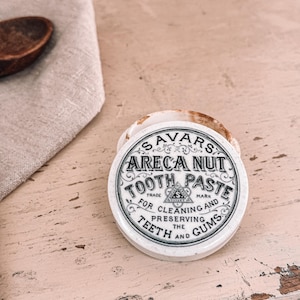 May include: A vintage white and brown Areca Nut Tooth Paste container. The lid features the text "SAVARS ARECA NUT TOOTH PASTE" in a decorative font. The container is open, revealing the inside. A wooden spoon and a linen cloth are in the background.