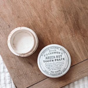 Puede incluir: Un tarro de cerámica blanca antigua con su tapa al lado, junto a una lata redonda de pasta dental Areca Nut de F.S. Cleaver. La lata tiene texto negro sobre fondo blanco, con "Tooth Paste" en un lugar destacado.