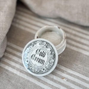 May include: An open, antique-style cold cream jar with a decorative lid. The lid features the words "Cold Cream" in a vintage font, surrounded by ornate floral and scrollwork designs. The jar is white with a slightly aged appearance, resting on a striped surface.