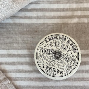 May include: A round, off-white vintage tin container featuring black decorative typography that reads S. MAW, SON & SONS CHERRY TOOTH PASTE FOR CLEANING & PRESERVING THE TEETH AND GUMS ALDERSGATE STREET LONDON ENGLAND with a central circular logo.
