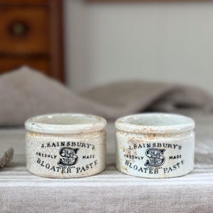Può includere: Due barattoli cilindrici in ceramica color bianco sporco con una finitura smaltata craquelé visibile. Ogni barattolo presenta una scritta nera che recita J. SAINSBURY'S FRESHLY MADE BLOATER PASTE e un logo S centrale con elementi floreali.