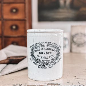 May include: A white ceramic jar with a cracked surface. The jar features a black and white label with the text "JAMES KEILLER & SONS DUNDEE MARMALADE" and "ONLY PRIZE MEDAL FOR MARMALADE LONDON, 1862". The jar is a vintage kitchenware item.