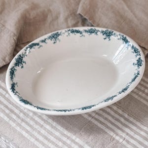 May include: Oval white serving dish with a teal floral border. The dish is set on a neutral-toned striped fabric. The floral design is a repeating pattern around the rim of the dish.