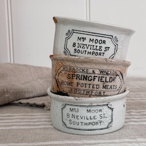 May include: A stack of three white ceramic pots with black printed text. The top and bottom pots are labeled Mrs Moor 8 Neville St Southport. The middle pot features a decorative border and reads Horrocks & Watson Springfield Home Potted Meats Southport.