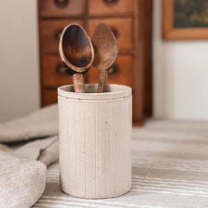 May include: A cream-colored, cylindrical utensil holder with vertical grooves, holding two wooden spoons. The spoons are dark brown with rounded bowls and long handles. The holder sits on a striped surface.