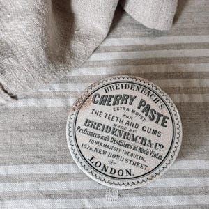 May include: A vintage, round tin of Breidenbach's Cherry Paste, with text in black and white. The tin is on a striped, neutral-toned fabric. The text reads "Extra Moist for the Teeth and Gums."
