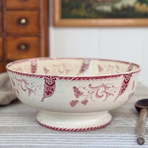 May include: Cream-colored pedestal bowl with a red floral pattern. The bowl has a wide, shallow design with a decorative rim and a sturdy base. The floral design includes stylized leaves and flowers, adding a vintage touch to the piece.