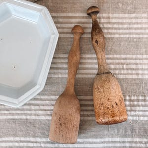 May include: Two wooden muddlers with rounded handles and rectangular heads. One is lighter in color, while the other is darker with visible wood grain. A white, octagonal dish is in the background. The items rest on a striped, neutral-toned surface.