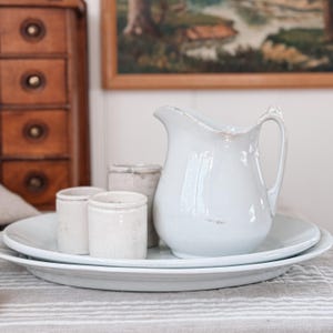 Puede incluir: Una jarra de cerámica blanca con asa, colocada sobre un plato blanco, acompañada de pequeños tarros de cerámica envejecida. Los objetos están dispuestos sobre un mantel a rayas, sugiriendo una estética vintage o rústica.