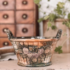 May include: A ceramic planter shaped like a basket with a woven pattern and floral accents. The planter has two handles and features raised roses and leaves.