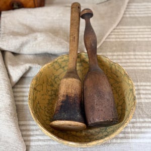 Könnte beinhalten: Eine gelbe Keramikschale mit grünem Craquelé-Muster, die zwei Holzstößel enthält. Ein Stößel ist hell mit einer dunklen, verbrannten Basis, während der andere aus dunklem, glattem Holz mit einem abgerundeten Griffende besteht.