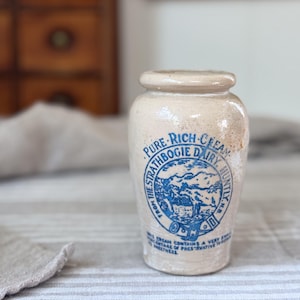 May include: A beige ceramic cream jar with a blue printed label that reads PURE RICH CREAM FROM THE STRATHBOGIE DAIRY HUNTLY N.B. The label features a circular graphic of a landscape with a cow and text about the cream's ingredients.