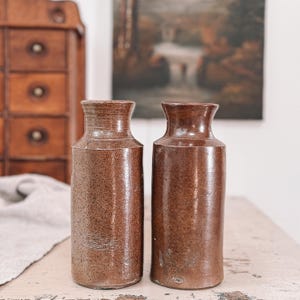 Puede incluir: Dos jarrones antiguos de gres marrón con superficie texturizada. Los jarrones tienen forma cilíndrica con cuello ligeramente acampanado. Los jarrones se exhiben sobre una mesa blanca envejecida.