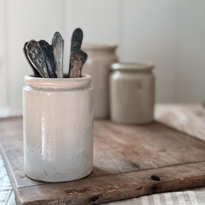 Può includere: Un vaso in gres beige chiaro con una collezione di posate d'epoca. Il vaso è appoggiato su un tagliere di legno. Altri due vasi in gres sono sullo sfondo. La scena ha un'estetica rustica e country.
