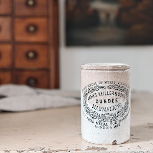 May include: A vintage Dundee Marmalade jar with a white and cream-colored exterior. The jar features black text and floral designs, including the words "James Keiller & Sons" and "Dundee Marmalade."