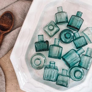 May include: A collection of small, antique teal glass bottles in various shapes, displayed in a white, hexagonal dish. A wooden spoon and a linen cloth are visible in the background.