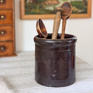 May include: A dark brown cylindrical stoneware crock with a thick rim, containing three wooden kitchen utensils. The crock has a smooth, glazed finish and a simple, sturdy design. The wooden spoons and tool have varying shades of brown wood grain.