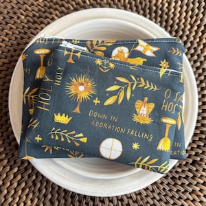 May include: Two dark teal fabric pouches with white zippers, featuring a repeating pattern of gold religious symbols and text. The pouches are resting on a white plate, with a woven brown background.