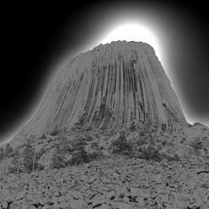 Puede incluir: Una fotografía en blanco y negro de una gran formación rocosa, conocida como Devils Tower, que se eleva desde el suelo. La roca es alta y delgada, con una superficie lisa y redondeada. El cielo está oscuro y nublado, y el sol brilla intensamente detrás de la roca.