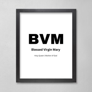 May include: Framed print with the text "BVM" in bold black letters, with "Blessed Virgin Mary" and "Holy Queen | Mother of God" below. The print is in a black frame and hangs on a white wall.