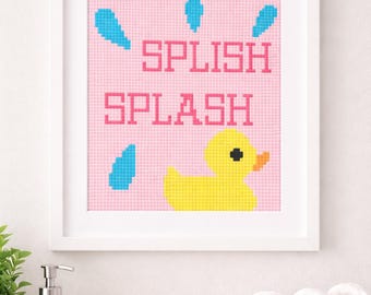 May include: A framed cross-stitch artwork with a pink background and the words "SPLISH SPLASH" in pink pixelated letters. Blue water droplets surround a yellow rubber duck. The artwork is in a white frame.