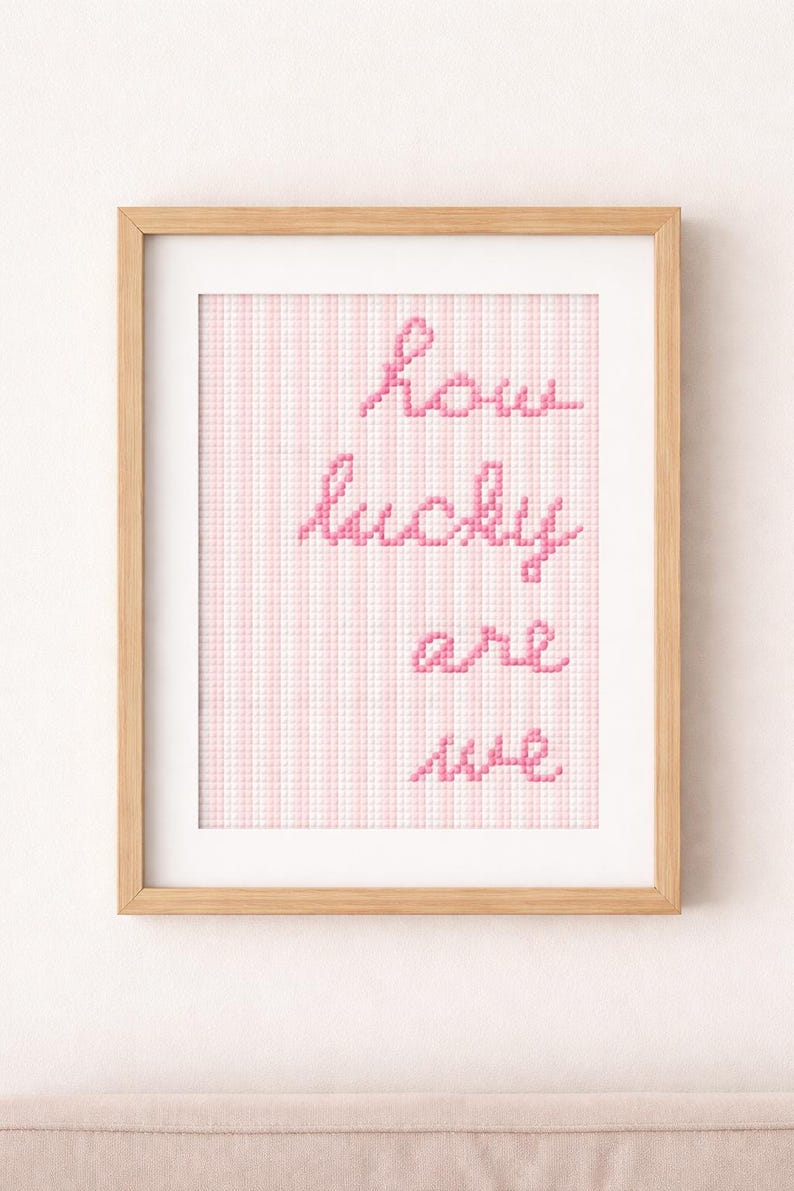 May include: A framed cross-stitch artwork with the words "how lucky are we" in pink thread. The background features a pink and white striped pattern. The frame is made of light-colored wood.