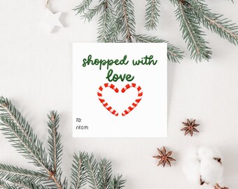 Op de afbeelding: Witte cadeaukaart met groene tekst die "shopped with love" zegt. Onder de tekst staat een rood-wit hart van snoepstokken. De kaart heeft onderaan de woorden "To:" en "From:".