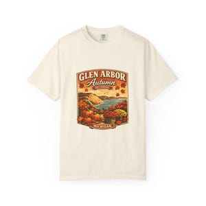 May include: A cream-colored short-sleeve t-shirt featuring a graphic print of a landscape with pumpkins, autumn leaves, and a lake. The text reads GLEN ARBOR Autumn FALL FESTIVAL MICHIGAN, framed in a decorative border with warm orange and red tones.