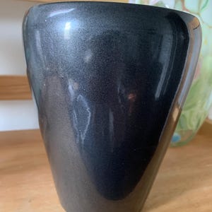 May include: A dark grey, metallic-finish vase with a flared, asymmetrical shape. The vase has a smooth, reflective surface and a wide opening. The background includes a glimpse of a colourful glass vase and a wooden surface.