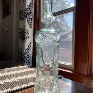 May include: A tall, clear glass bottle with a light green tint, featuring a slender neck topped with a cork stopper. The bottle has a cylindrical body with textured, embossed leaf patterns around the top and bottom sections of the main glass structure.