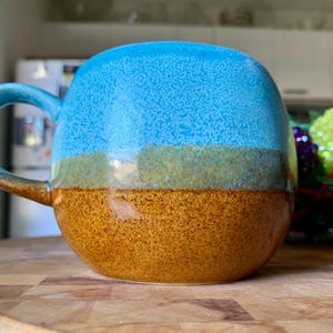 May include: A round ceramic mug with a blue top, a green-brown middle band, and a textured brown bottom. The mug has a matching blue handle. The mug is sitting on a wooden surface.