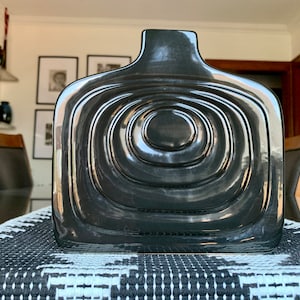 May include: A dark gray, square-shaped vase with a glossy finish. The vase features a series of concentric, circular ridges that create a textured surface. The vase is placed on a black and white patterned table runner.