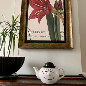 May include: White teapot with a black hat and a smiling face. The teapot has red accents on the face and a black spout. The teapot is sitting on a wooden surface.