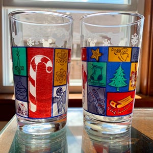 May include: Two clear glass tumblers with a Christmas theme. The glasses feature a colorful patchwork design with Mickey Mouse icons and snowflakes. The design includes a candy cane, a Christmas tree, a stocking, and a sled.
