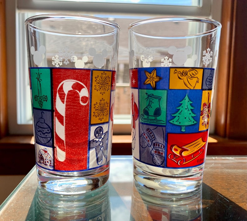 May include: Two clear glass tumblers with a Christmas theme. The glasses feature a colorful patchwork design with Mickey Mouse icons and snowflakes. The design includes a candy cane, a Christmas tree, a stocking, and a sled.