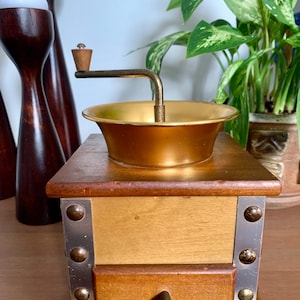 Könnte beinhalten: Eine Vintage-Kaffeemühle mit Holzsockel und Messingschüssel und -griff. Die Mühle hat eine Schublade und Metallakzente. Das Design ist klassisch und das Holz hat eine warme, natürliche Oberfläche. Ein dekorativer Artikel für die Küche.