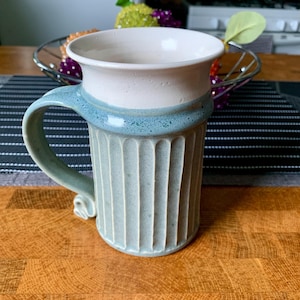 May include: A ceramic mug with a white top and a light green bottom. The mug has a fluted design and a handle.