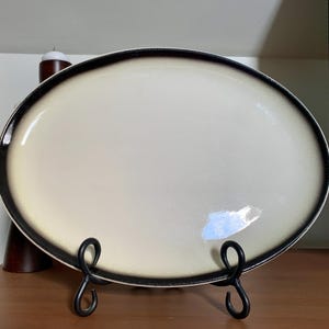 May include: An oval serving platter with a cream-colored center and a dark brown rim. The platter is displayed on a black metal stand with a scrolled design. The platter is likely ceramic or porcelain.