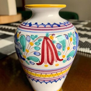May include: A white ceramic vase featuring a hand-painted floral pattern in red, teal, and blue. The vase has a yellow rim and base, with horizontal purple and blue decorative bands encircling the body, creating a colorful and symmetrical design.