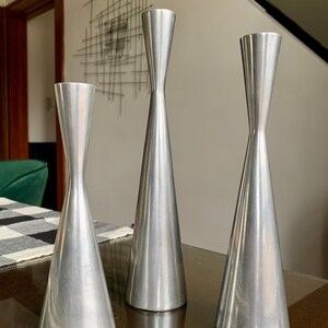 May include: Three silver-colored candle holders with a conical base and a slender, elongated neck. The candle holders are displayed on a reflective surface. The design is minimalist and modern, suitable for home decor.