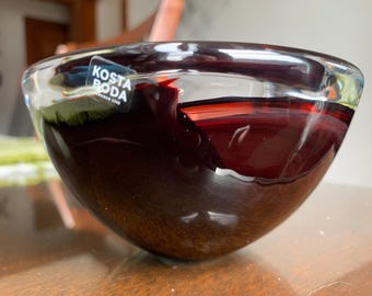 May include: A clear glass bowl with a dark red and brown swirl design. The bowl has a sticker on the side that says "Kosta Boda".