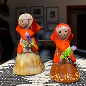May include: Two ceramic figurines of women wearing red hooded dresses and holding bouquets of purple flowers. The figurines are standing on a black and white patterned surface.