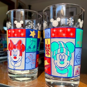 May include: Two clear glass drinking glasses with a Christmas theme. One glass features Mickey Mouse wearing a green hat and scarf, the other features Minnie Mouse wearing a red bow. Both glasses have a patchwork design with various Christmas symbols.