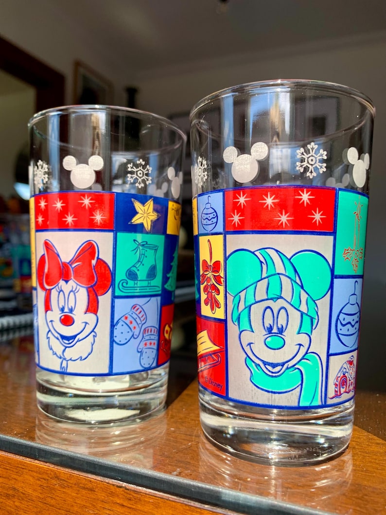 May include: Two clear glass drinking glasses with a Christmas theme. One glass features Mickey Mouse wearing a green hat and scarf, the other features Minnie Mouse wearing a red bow. Both glasses have a patchwork design with various Christmas symbols.