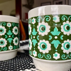 May include: Two white ceramic mugs with a green and white floral pattern. The mugs have a wide base and a slightly flared top.